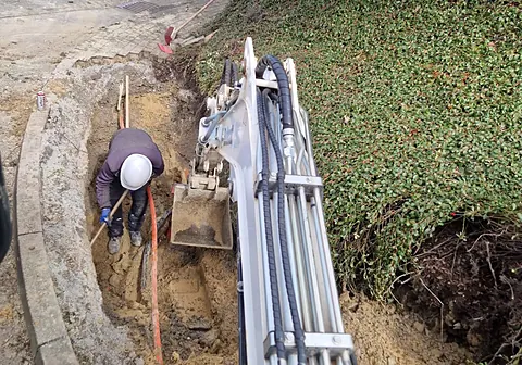 Monteur mit Helm im Graben am Bordstein, Minibagger daneben
